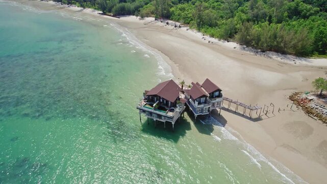 Cinematic drone orbit around an abandoned overwater resort villa showing visible deterioration and structural damage beside turquoise tropical waters and a sandy beach in Malaysia.