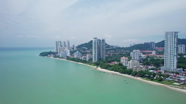 Wide aerial establishing shot of coastal Penang, Malaysia, featuring high-rise residential development, beachfront communities, tropical water and densely developed hillsides.
