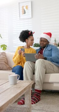 Vertical video: Diverse couple with credit card on sofa scrolling app on tablet for gift shopping