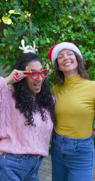 Vertical video: Two adult women friends raising candy canes adjusting Santa hat laughing on yard