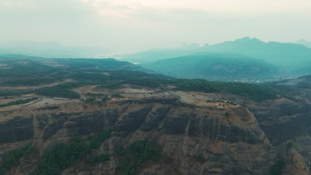 Aerial view of Tiger Point plateau reveals layered mountain ranges and valley settlements at sunrise in Lonavla, Maharashtra, India. Atmospheric haze creates depth across the Western Ghats.