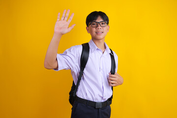 Indonesian Schoolboy with Backpack and Glasses Waving Hand with Open Palm, Yellow Background © Gatot