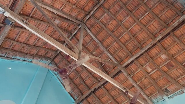A ceiling fan begins to rotate beneath a traditional terracotta tile roof in a rural Indian school. The wooden beam structure and blue painted walls frame the spinning fan blades.