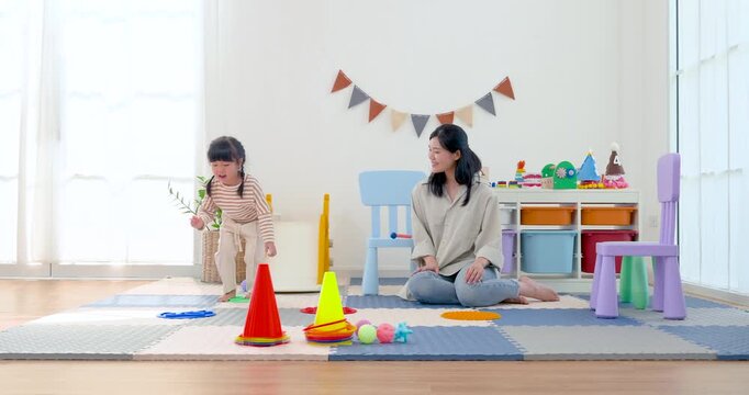 Asian mom kneels on play mat as preschool daughter moves between cone markers, building coordination, confidence, body awareness, and playful gross motor development through guided indoor activity.