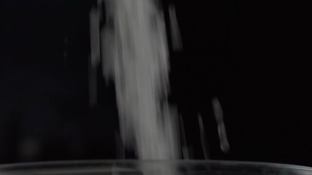 Fine white sugar pours steadily into dark container, forming a flowing stream of granules against black background during food preparation
