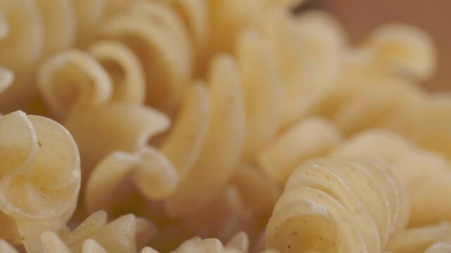 Macro close-up shows uncooked fusilli pasta with curved spiral shapes and ridged surfaces, highlighting pale golden color, dry texture, and detailed pasta forms.