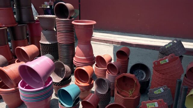 Colorful plastic and terracotta plant pots are displayed in stacks and rows at an outdoor market, showing various shapes, sizes, and earthy tones under bright sunlight.
