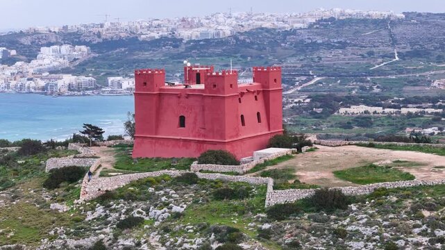 red fortress tower close orbit hilltop crenellations visitors walking grounds distant island across channel coastal bay salt pans malta aerial drone footage historic watchtower landmark