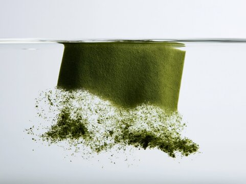A piece of green powder dissolves in water, creating a cloud of particles and representing the scientific concept of dissolution and dispersion.