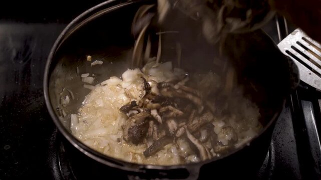Adding fresh sliced shiitake mushrooms to sauteed onions in a cast iron pot on kitchen stove.