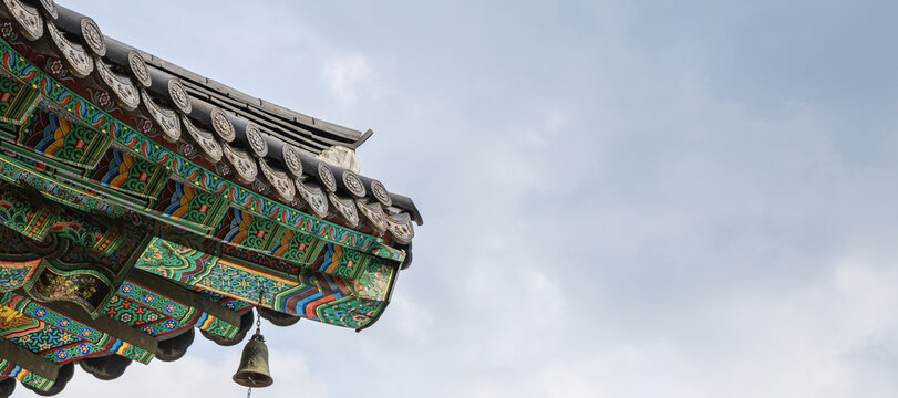 Ornate double eaves with colorful dancheong paint and wind chime on tiled roof of traditional Korean Buddhist temple against sky with copy space.