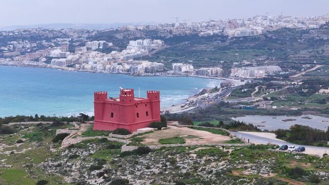 red tower crenellated watchtower stone battlements hilltop visitor pathway parked cars overlooking sandy beach turquoise mellieha bay coastal town skyline malta aerial drone footage clifftop landmark
