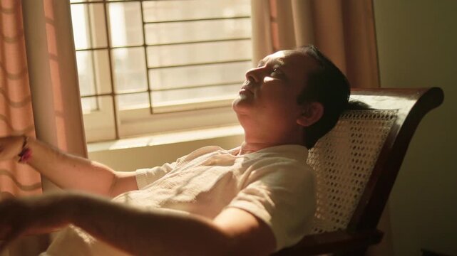 Senior Indian man resting comfortably in an armchair beside a bright window at home.