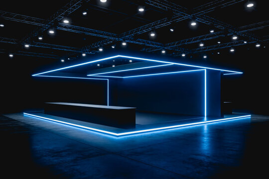 A modern empty exhibition booth with blue neon lighting, set in a dark indoor event venue with ceiling trusses and spotlights.