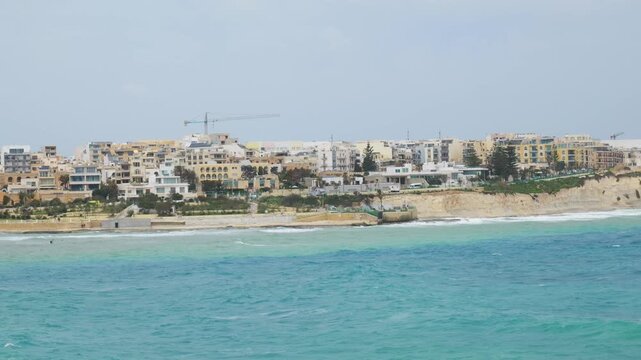 apartment buildings coastal town limestone bluff turquoise sea construction crane mediterranean skyline malta coast waterfront development cloudy sky aerial view