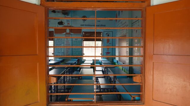 An empty classroom with rows of wooden desks and benches is viewed through an orange-painted window frame with metal grille bars. The room features blue walls, educational posters, and natural.