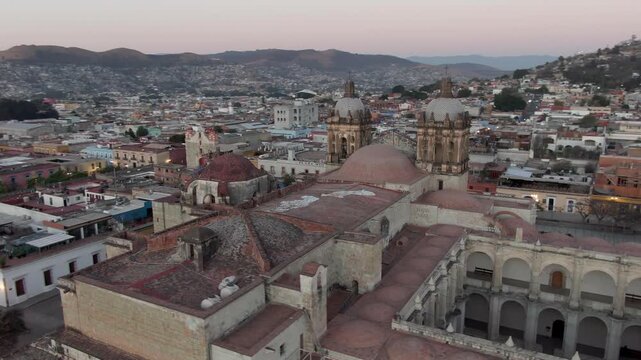 Aerial drone shot of historic cathedral complex with domes, towers, courtyards and weathered stone architecture surrounded by dense urban neighborhoods in Oaxaca, Mexico