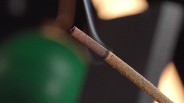 Close up macro shot of burning incense stick releasing smoke against dark blurred background, highlighting textured wood, glowing tip and atmospheric detail.