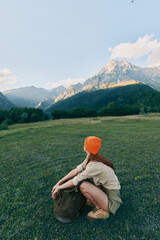 Naklejka premium Woman sits in a wide grassy field with a backpack and orange beanie, enjoying a peaceful outdoor lifestyle amid distant mountains and expansive nature scenery.