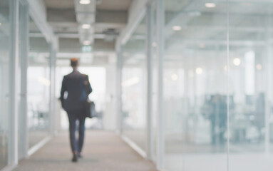 Obraz premium Professional worker walking inside a modern office surrounded by glass partitions and bright work areas. The blurred environment provides a clean background for advertising and business content.
