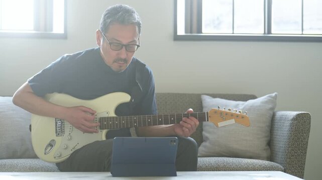 タブレットを見ながらエレキギターを弾く男性。音楽、楽器、演奏、練習、趣味、動画、バンド、音楽家、作曲