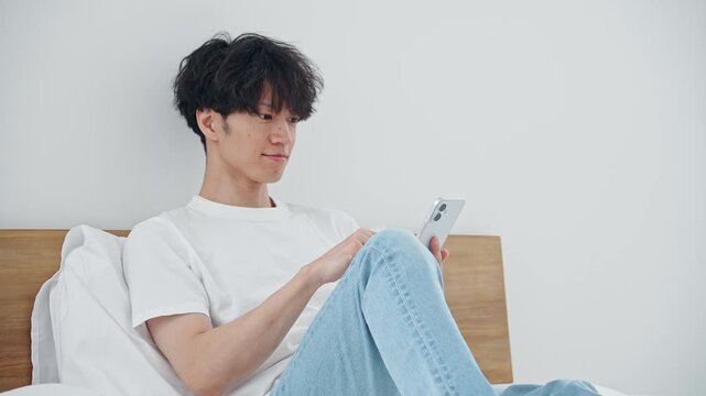 寝室でスマホを使う男性。アプリ、SNS、動画、リラックス、ベッド、ゲーム、笑顔、検索、AI、DX