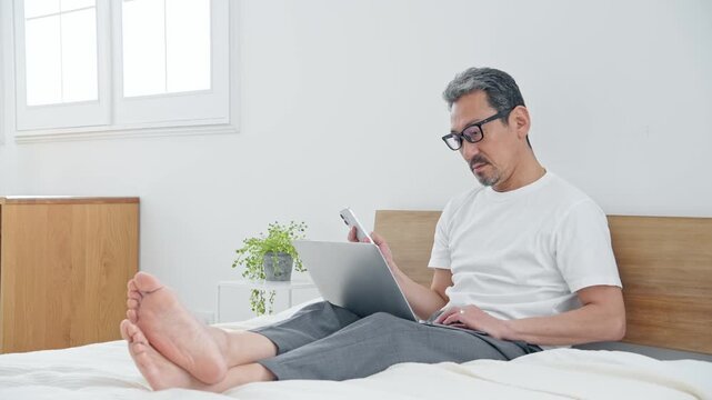 ベッドでノートPCを使うシニア男性。パソコン、仕事、DX、スマホ、アプリ、ワーケーション、テレワーク
