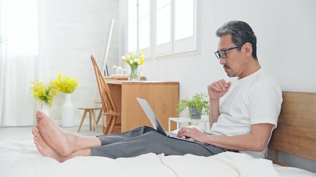 ベッドでノートPCを使うシニア男性。パソコン、仕事、DX、リラックス、ワーケーション、テレワーク