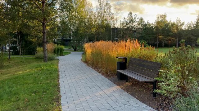 Alley with pedestrian path made of paving slabs and benches in residential complex park. Bench in garden path urban spaces. Walkway in city park at modern buildings. Serene path winds through park 