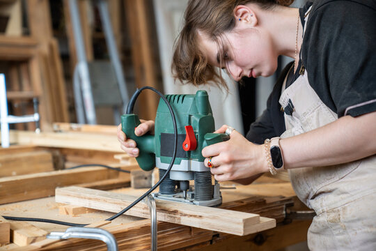 Focused skillful woman restorer working with electric plunge router precisely cutting and trimming surface on wooden detail, female crafter carefully processing material with handheld wood router.