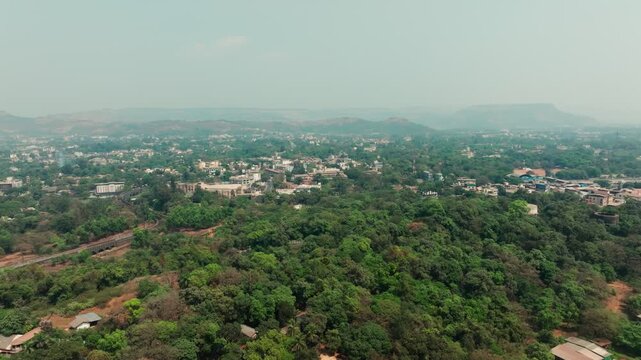 Lonavla town spreads across hilly terrain with dense green forest covering the foreground and residential buildings visible beyond. Misty hills fade into the horizon under overcast skies.