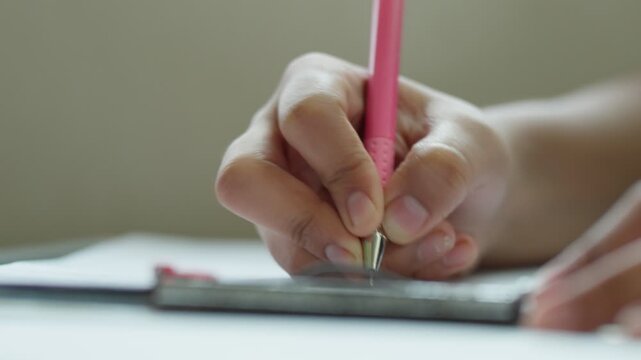 Close-up of a student's hand gripping a pink pen while writing in a notebook on a desk in school in India. The camera remains static, focused on the hand and writing motion in shallow depth of field.