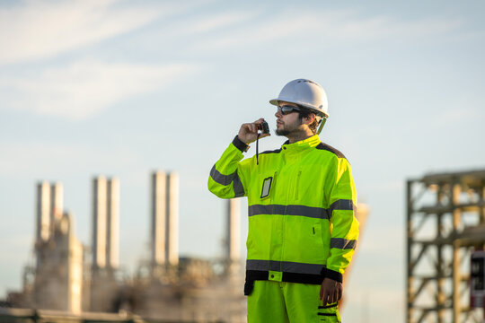 Oil refinery engineer using walkie talkie at industrial plant, petroleum plant worker in safety gear talking on radio outdoor, energy industry professional communicating with walkie talkie at facility