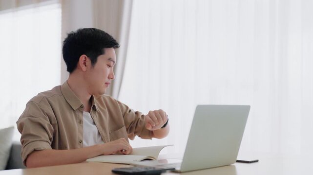 Happy young Asian man working on laptop in living room. Freelance worker male sitting at desk writing notes while watching webinar, studying online