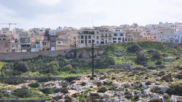 cross monument hilltop overlooking coastal town aerial view drone footage rocky terrain green shrubs boundary wall malta apartment buildings rooftops construction crane religious memorial landmark