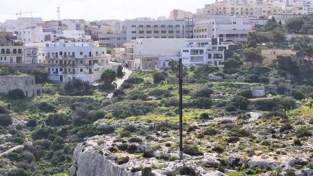 cross monument rocky hillside coastal town skyline aerial view drone footage green shrubs boundary wall malta apartment buildings rooftops narrow streets parked cars religious memorial landmark