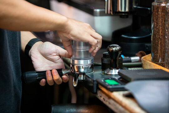 Selective focus to barista carefully pouring freshly ground coffee powder from a clear dosing cup into a metal portafilter at a cafe counter.