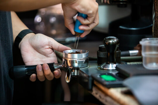 Selective focus to barista using a needle distribution tool (WDT) to stir and break up clumps of ground coffee in a portafilter at a cafe counter.