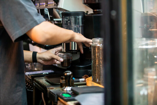Selective focus to Side view of a barista using an electric coffee grinder machine to catch freshly ground coffee powder into a cup at a cafe counter.
