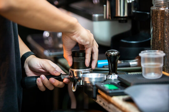 Selective focus to barista using a black coffee distributor leveler tool in a portafilter to evenly prepare ground coffee before tamping at a cafe counter.