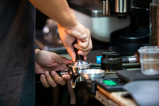 Selective focus to professional barista using a hand tamper to press ground coffee powder in a portafilter at a coffee shop counter to prepare fresh espresso.