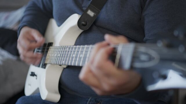 Close up shot of a person playing chords on a white electric guitar while practicing music at home.