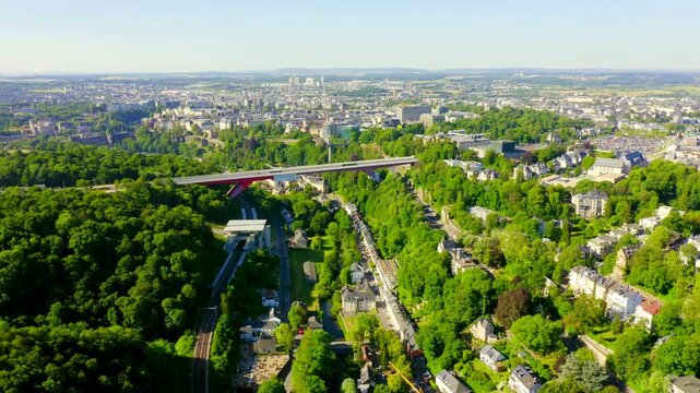 Luxembourg. The vivid red steel span of Pont Rouge cutting across the lush Alzette valley, Drone footage over dense green canopy with the historic city center in the distance under clear morning ligh