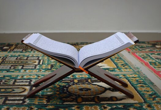 Open Quran on a wooden stand placed or rakel on a patterned prayer mat, representing Islamic faith, spirituality, prayer, worship, religious education, and peaceful Muslim lifestyle.