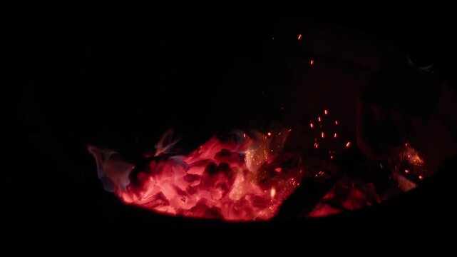 Slow motion of glowing red coal and sparks flying in dark fire pit.