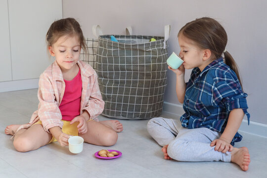Two Young Girls Sit Cross Legged On Floor With Small Cups And Snack Plate Near Woven Storage Basket In Bright Minimal Room, Showing Quiet Home Playtime, Friendship