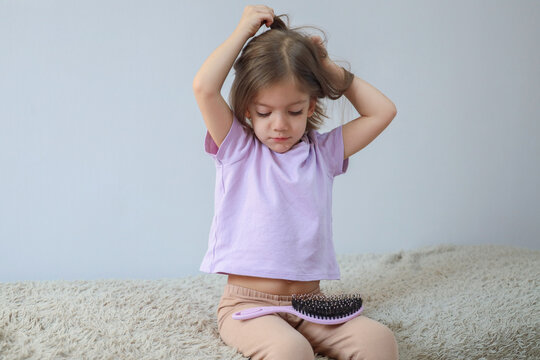 Young girl sitting on soft rug combing hair with pink brush, wearing lilac shirt and beige pants, calm indoor grooming moment against plain light background, with girl, child, hair