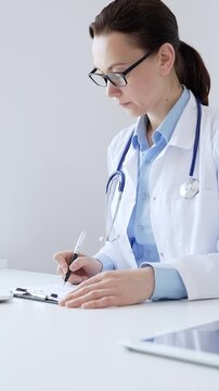 Middle aged female doctor completing healthcare paperwork, clipboard positioned near laptop and stethoscope in well lit clinical workspace by window. Medicine and health care