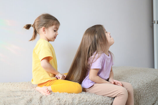 Two young girls sitting on soft shag carpet indoors while one gently plays with other girl's long hair, colorful casual clothes, bright natural light, calm home scene