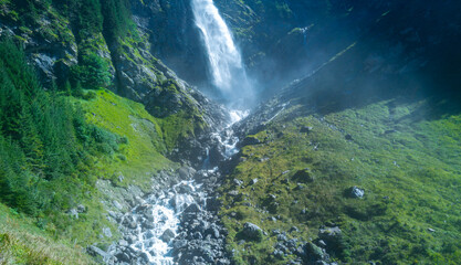 Naklejka premium Waterfall cuts through a rocky gorge. Waterfall rushes down dark alpine rocks. Mountain landscape contains waterfall inside wilderness. Swiss nature surrounds waterfall near gorge.